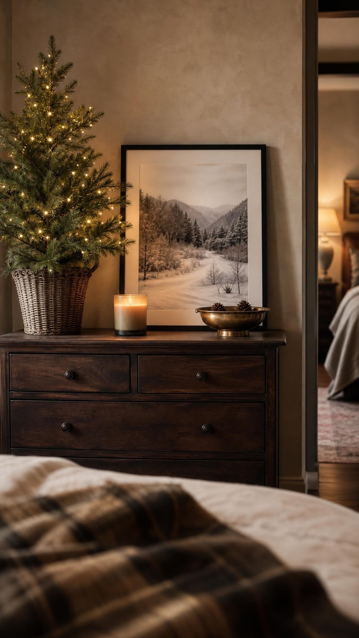 A styled dresser vignette with a small lit tree, a framed winter landscape print, a candle and a brass dish, neutral bedroom, warm light