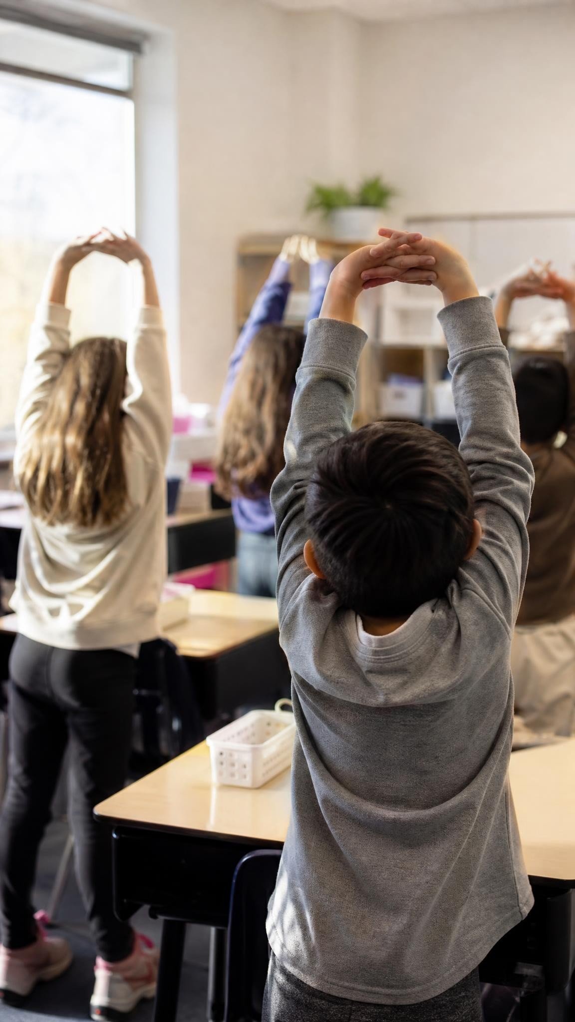 Elementary children seen from behind stretching their arms high above their heads beside their desks in a bright classroom