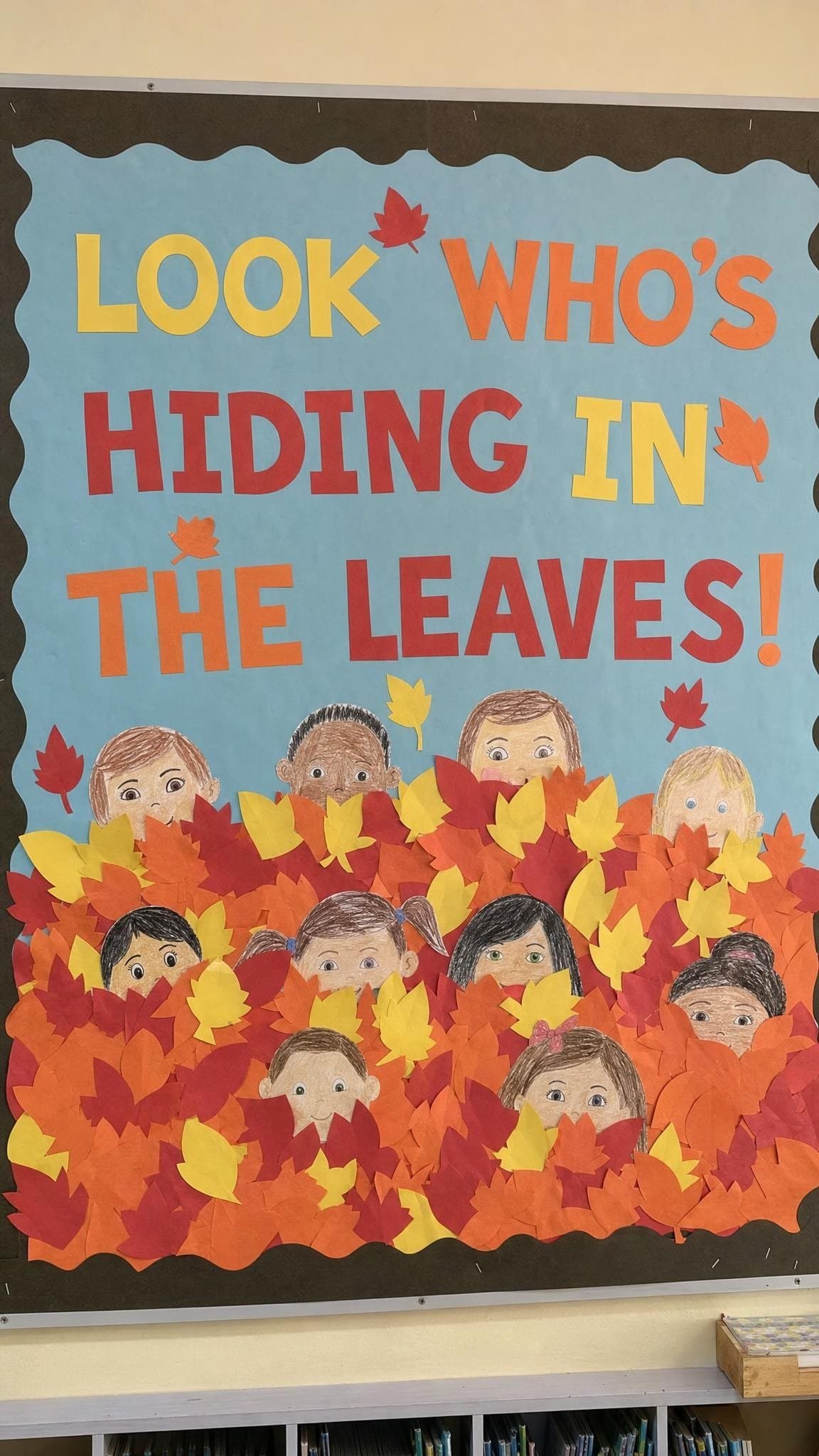 October bulletin board with children's self portraits peeking out of a pile of paper leaves
